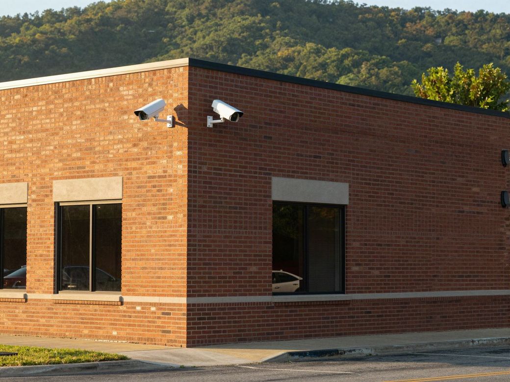 Brick commercial building with two security cameras on the corner and wooded hills behind